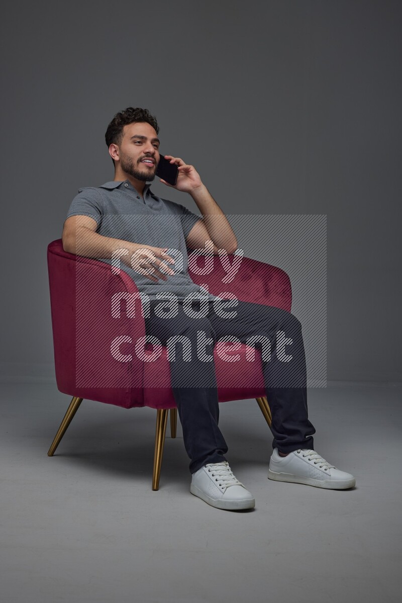 A man wearing casual and talking in his phone while setting on a burgundy chair eye level on a gray background