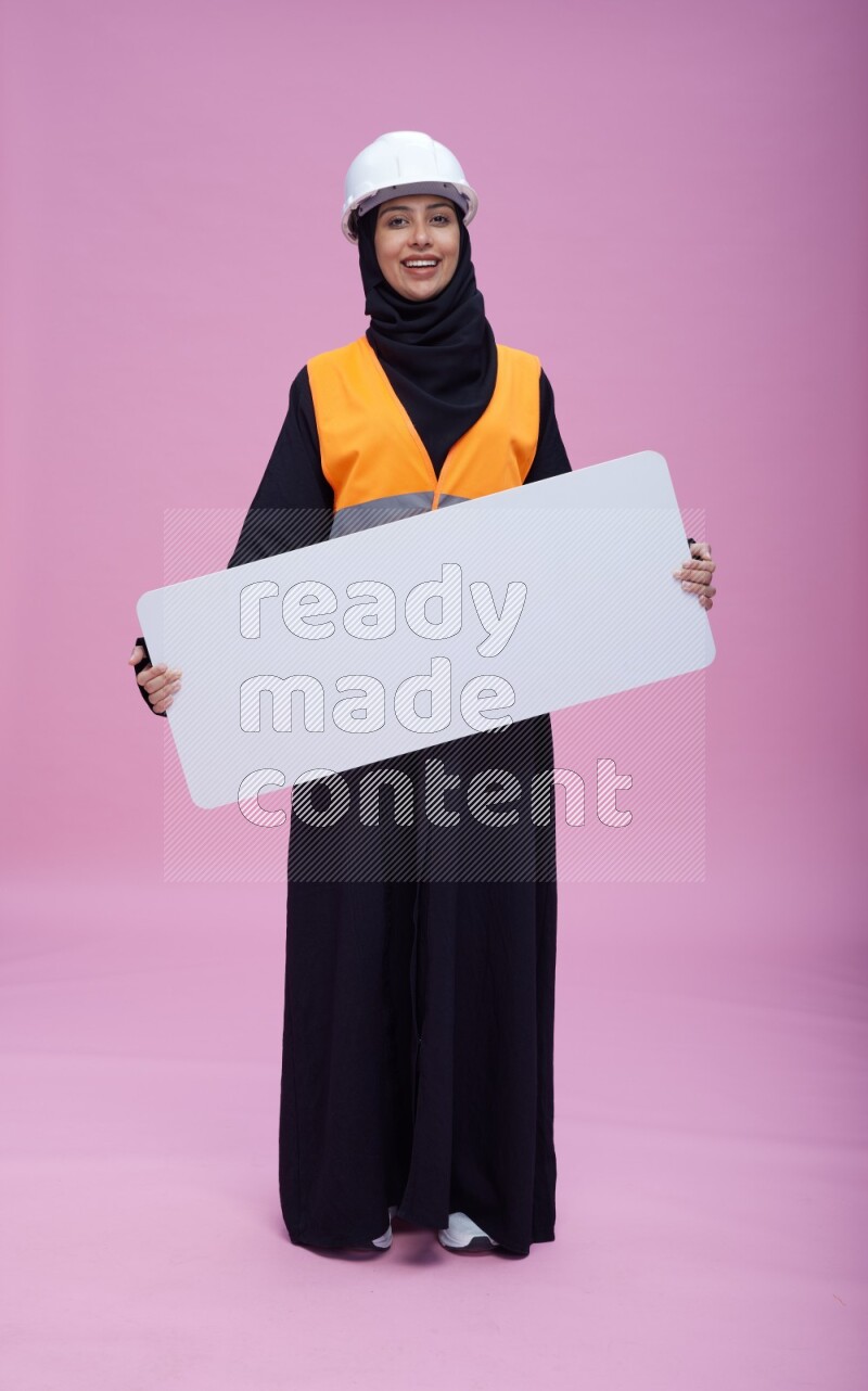 Saudi woman wearing Abaya with engineer vest and helmet standing holding board on pink background