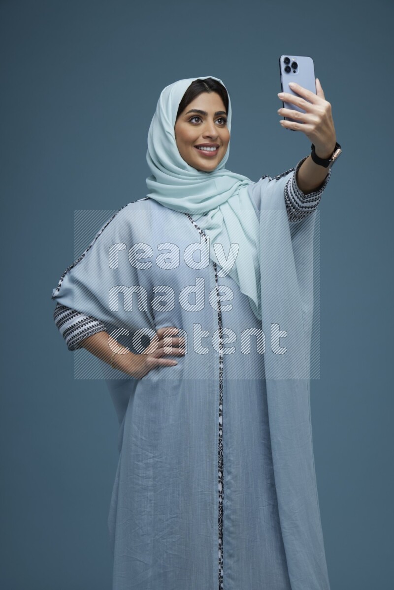 A Saudi woman Taking a Selfie on a blue background wearing a blue Abaya with hijab