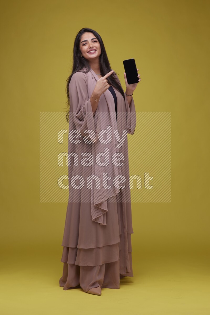 A woman Showing SmartPhone Screen on a Yellow Background wearing Brown Abaya