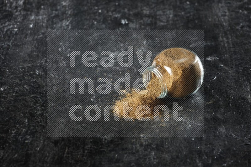 A flipped glass spice jar full of cumin powder and powder spilled out on a textured black flooring