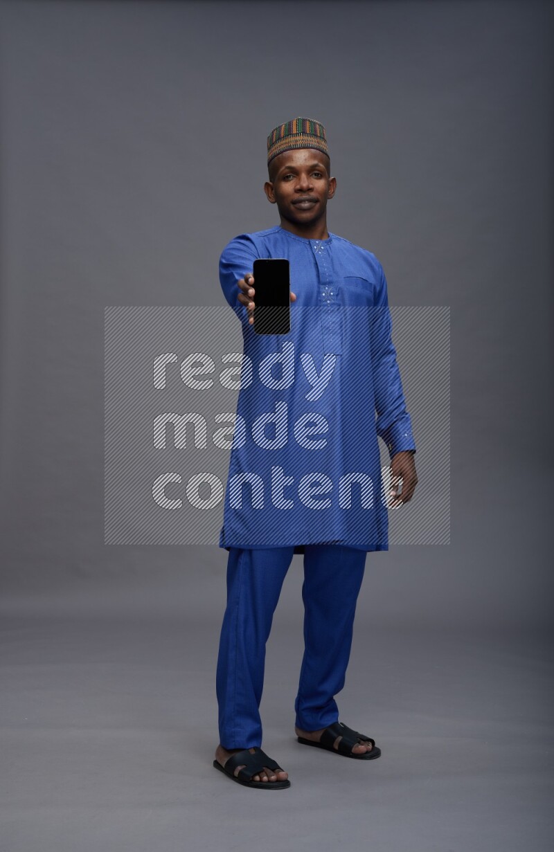 Man wearing Nigerian outfit standing showing phone to camera on gray background