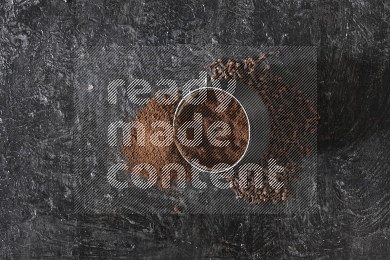 A black pottery bowl full of cloves powder spilled out of it with whole grains on a textured black flooring