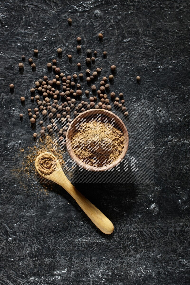 A wooden bowl and spoon full of allspice powder and whole balls spreaded on a black flooring