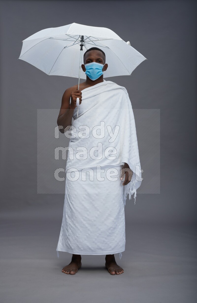 A man wearing Ehram with face mask Standing holding umbrella on gray background