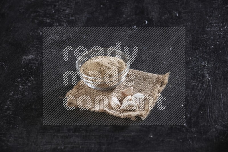 A glass bowl full of garlic powder placed on burlap fabric with garlic cloves on a textured black flooring