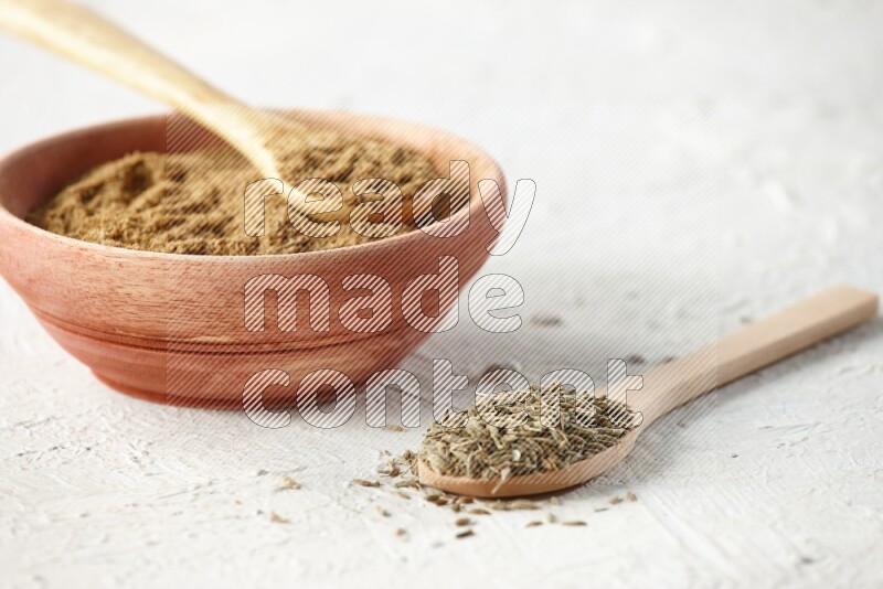 A wooden bowl and 2 wooden spoons full of cumin powder and cumin seeds on textured white flooring