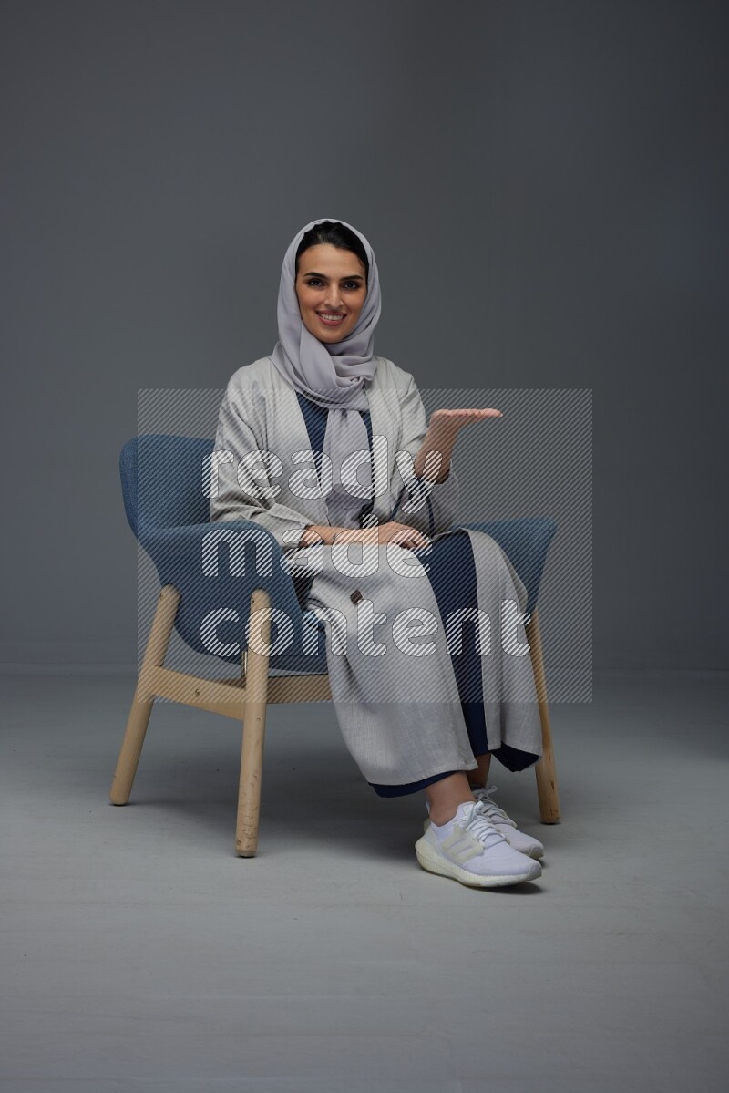 A Saudi woman wearing a light gray Abaya and head scarf sitting on a dark grey chair while making multi hand gestures eye level on a grey background