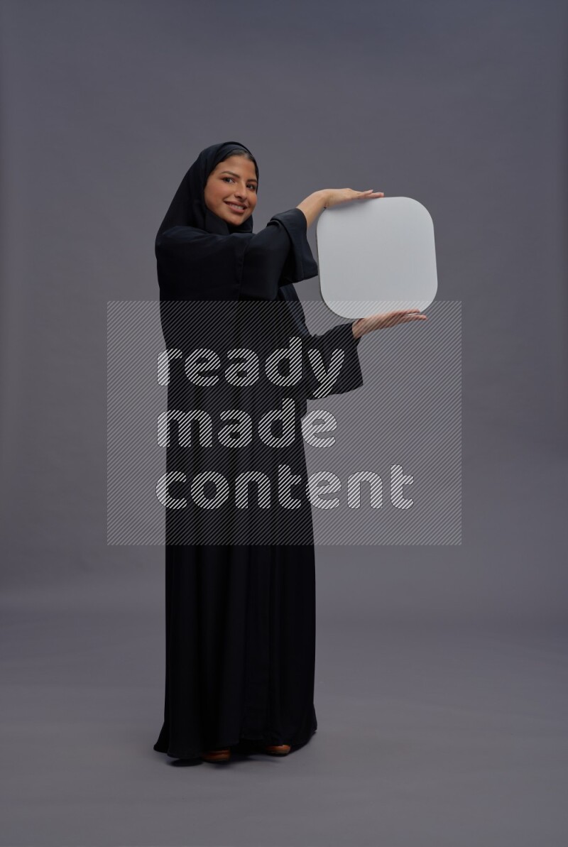 Saudi woman wearing Abaya standing holding social media sign on gray background
