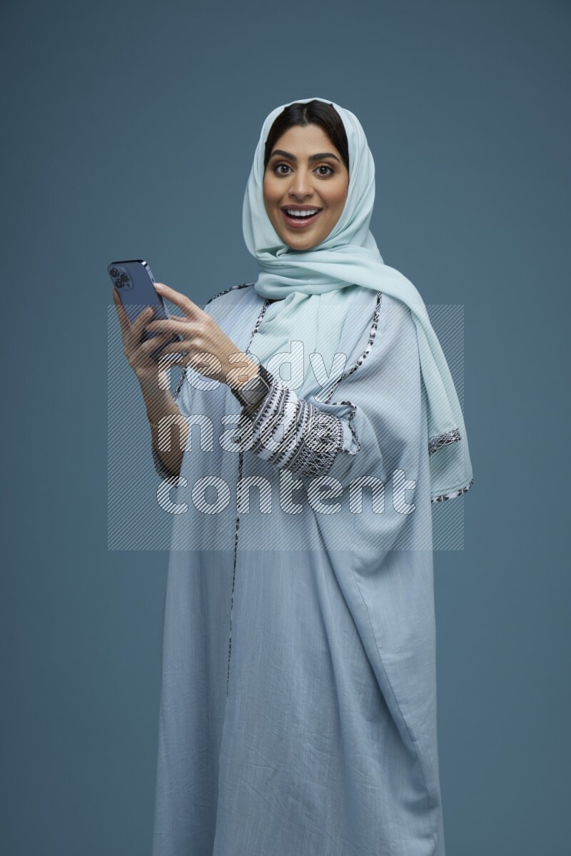 A Saudi woman posing with her phone in a blue background wearing a blue Abaya with hijab