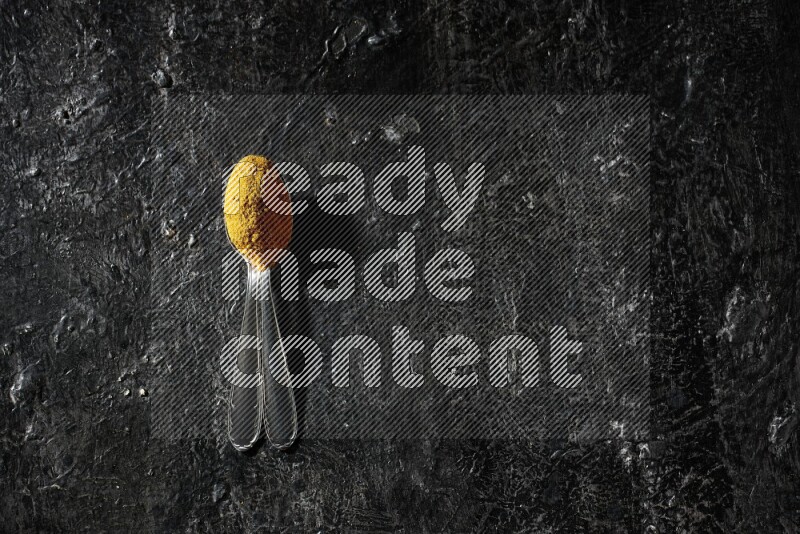 2 metal spoons full of turmeric powder on a textured black background