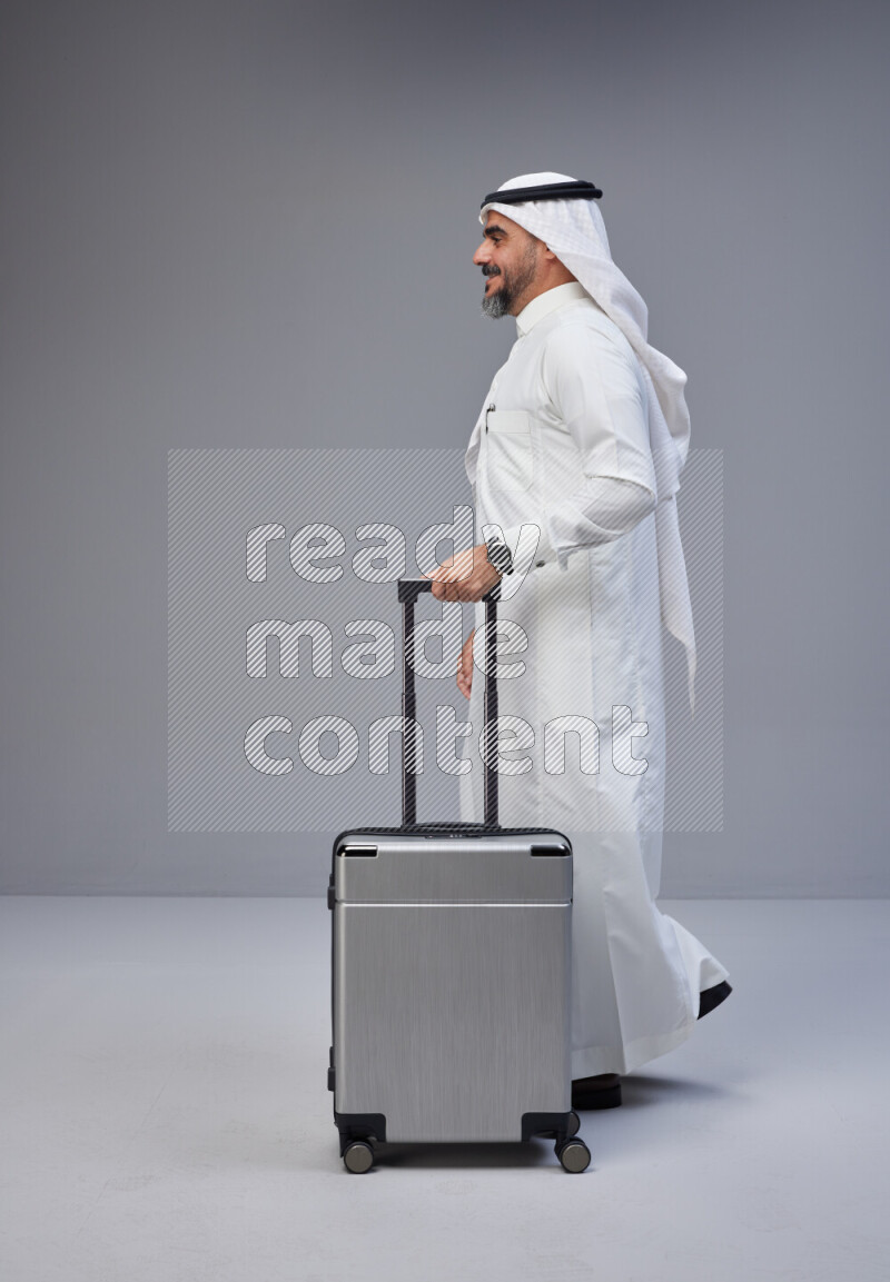 Saudi man wearing Thob and white Shomag standing holding Travel bag on Gray background