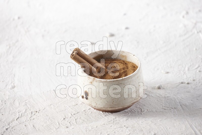 Ceramic beige bowl full of cinnamon powder with a cinnamon stick on a textured white background