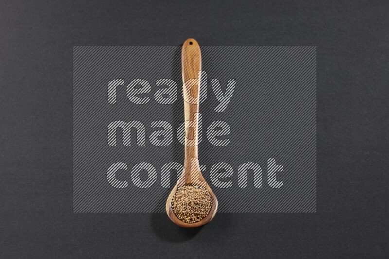 A wooden spoon full of mustard seeds on a black flooring