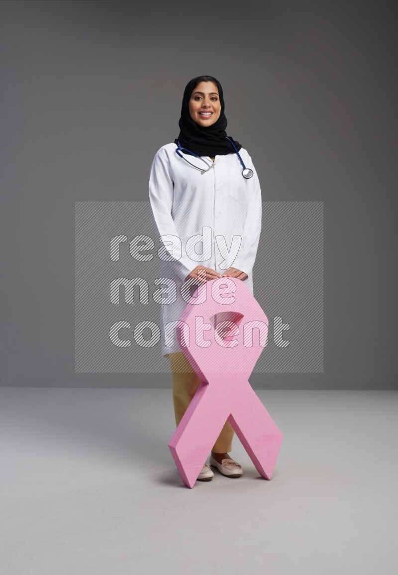 Saudi woman wearing lab coat with stethoscope standing holding awareness ribbon symbol standing on Gray background