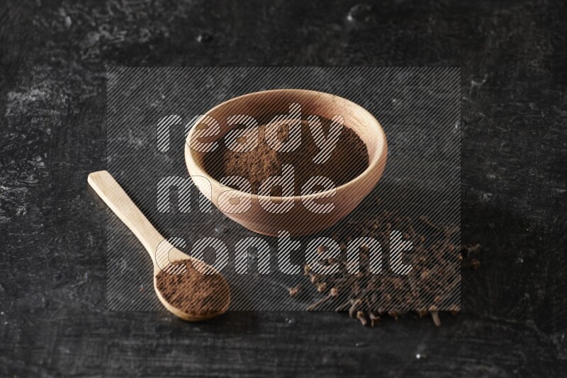 A wooden bowl and a wooden spoon full of cloves powder with spreaded cloves on a textured black flooring