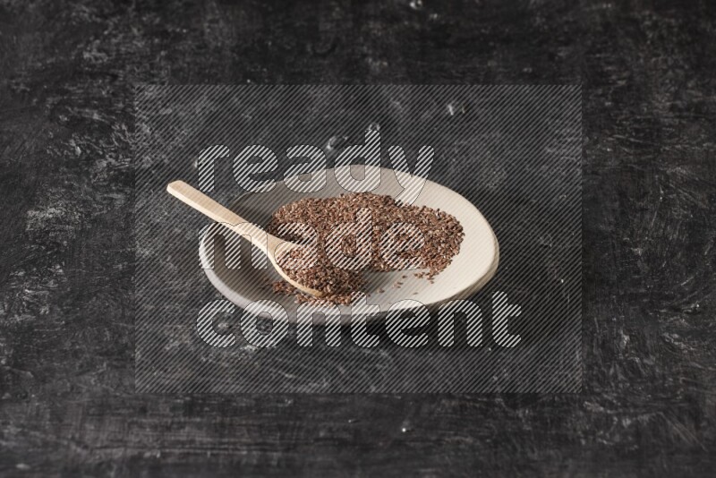 A multicolored pottery plate full of flaxseeds and wooden spoon full of seeds on a textured black flooring