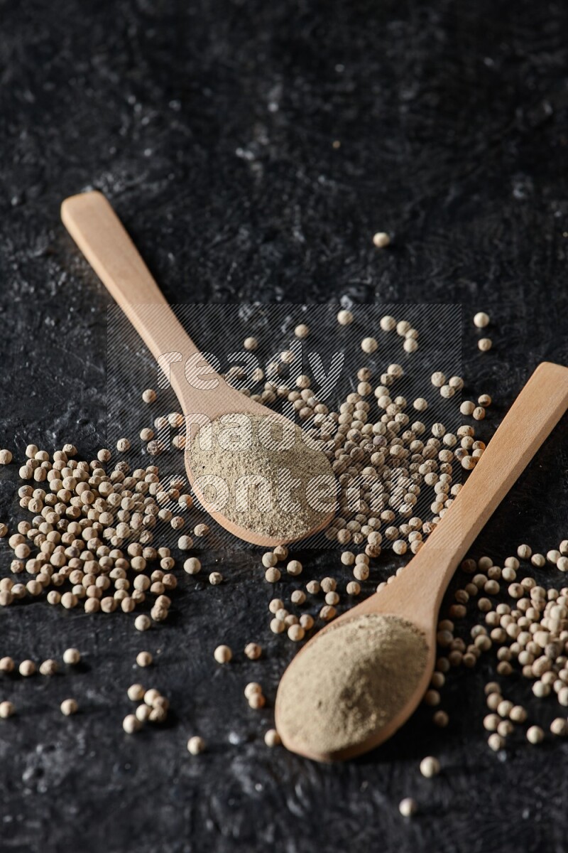2 wooden spoons full of white pepper powder with white pepper beads on textured black flooring