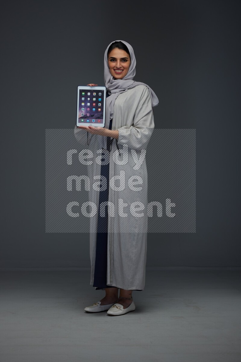 A Saudi woman wearing a light gray Abaya and head scarf standing and showing the phone's screen eye level on a grey background