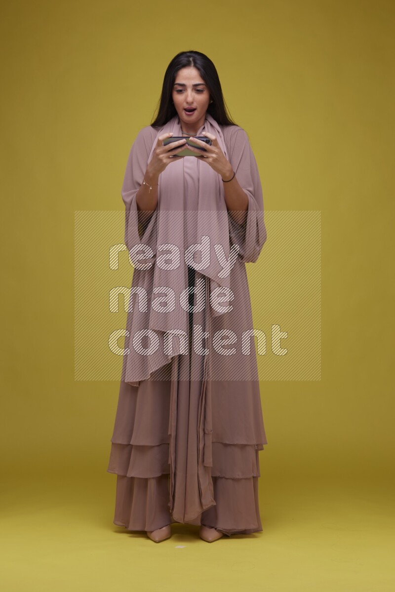 A woman Playing a Game on her smartphone on a Yellow Background wearing Brown Abaya