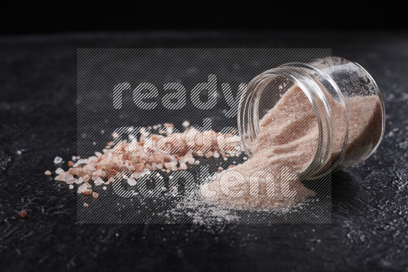 A glass jar full of fine himalayan salt with some himalayan crystals beside it on a black background