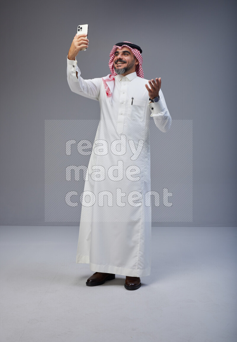 Saudi man Wearing Thob and red Shomag standing taking selfie on Gray background