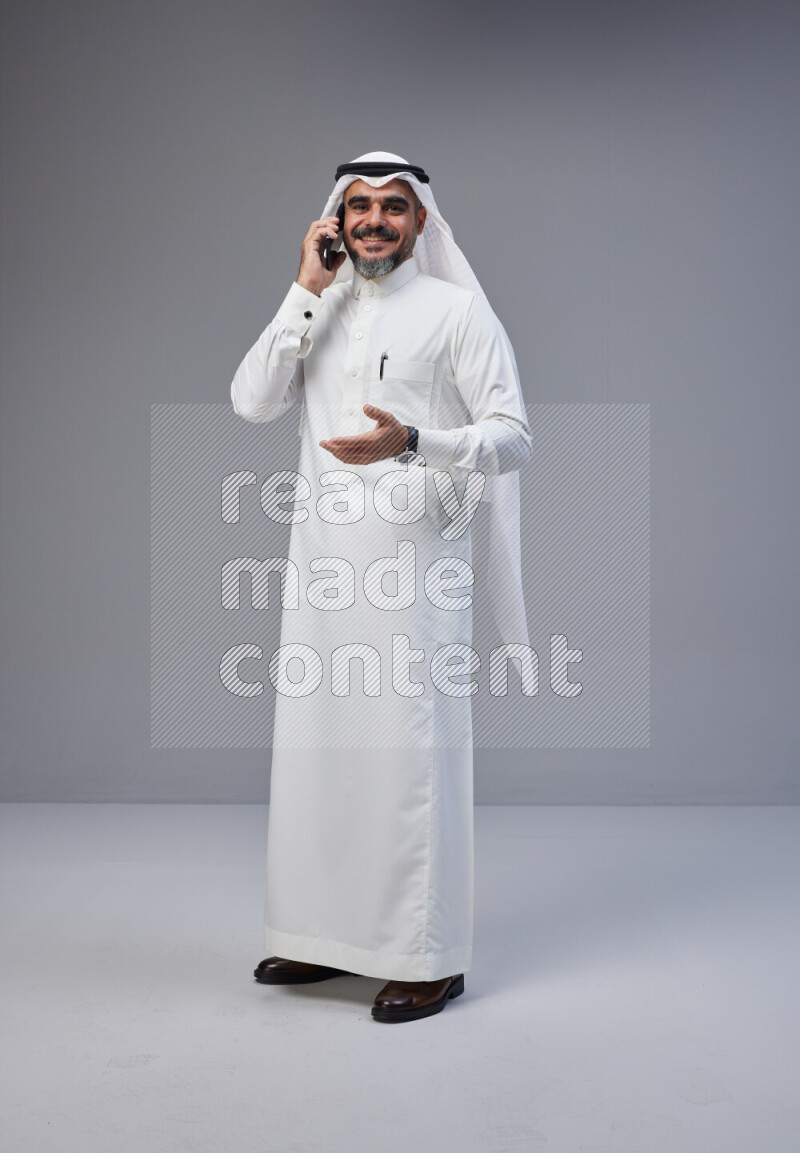 Saudi man Wearing Thob and white Shomag standing talking on phone on Gray background