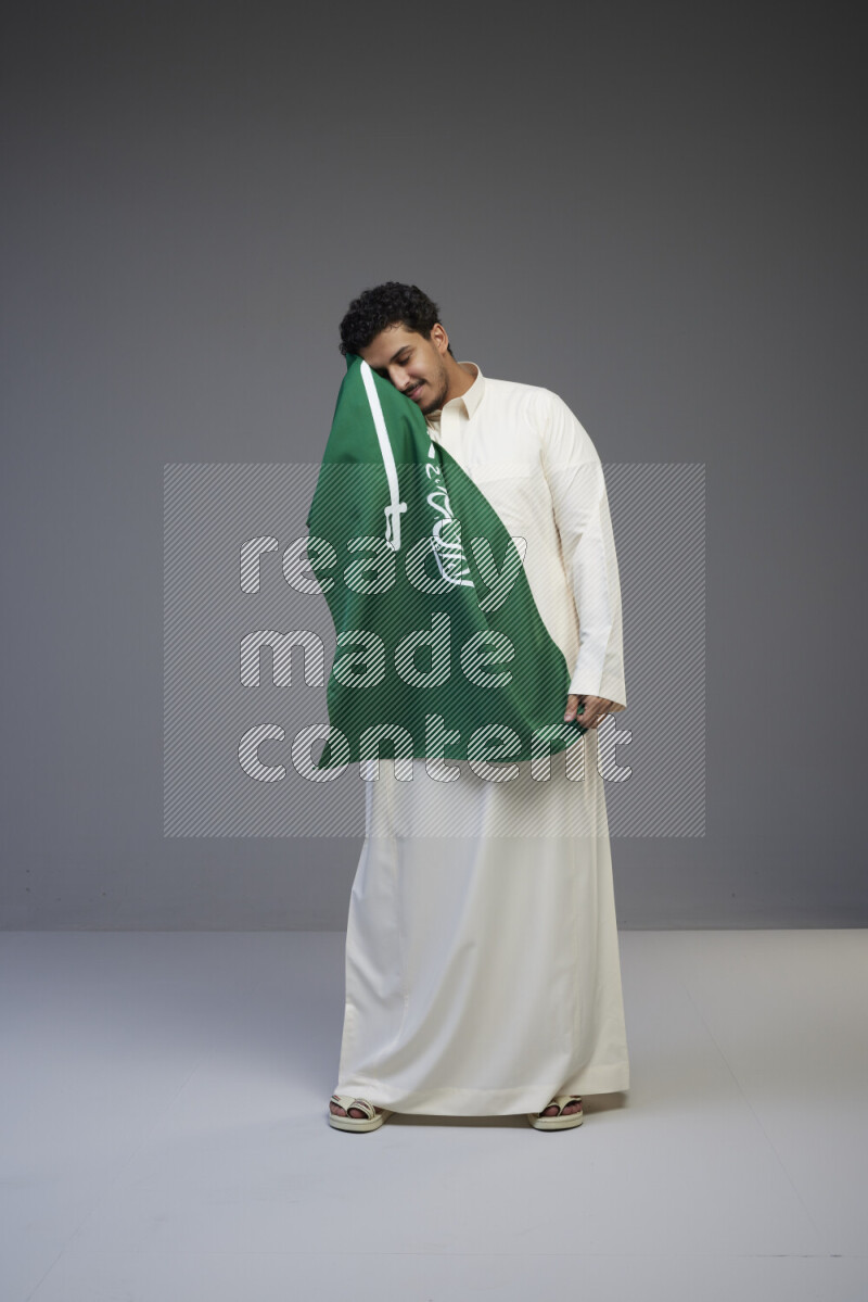 A Saudi man standing wearing thob holding big Saudi flag on gray background