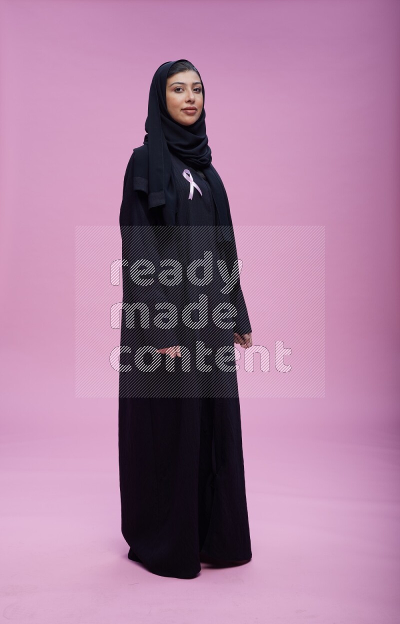 Saudi woman wearing pink ribbon on Abaya standing interacting with the camera on pink background