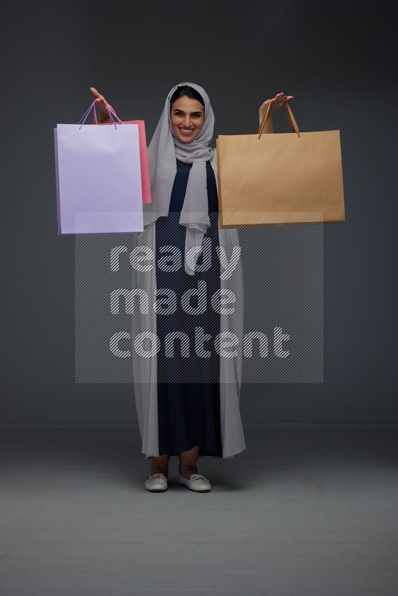 A Saudi woman wearing a light gray Abaya and head scarf standing and holding shopping bags on a grey background