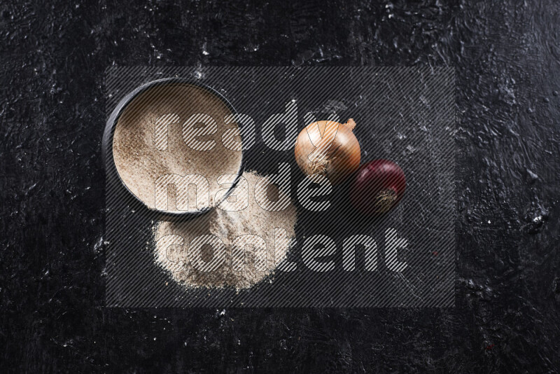 A black pottery bowl full of onion powder with fallen powder from it on black background