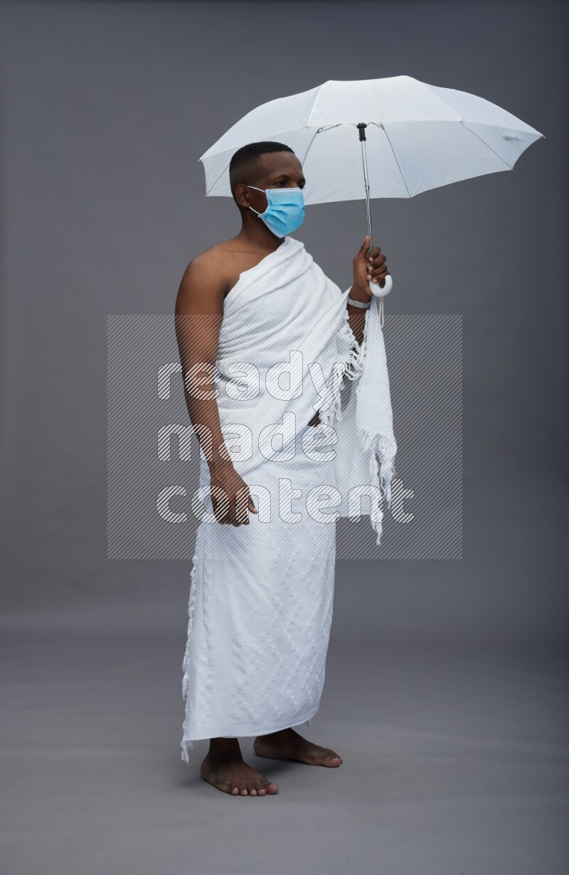 A man wearing Ehram with face mask Standing holding umbrella on gray background