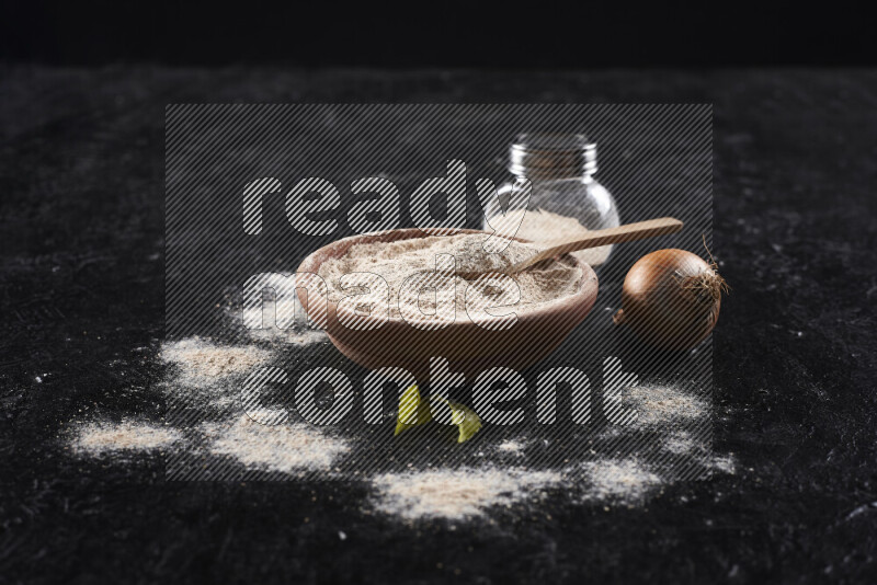 A wooden bowl full of onion powder with a glass jar beside it and fresh onion on black background