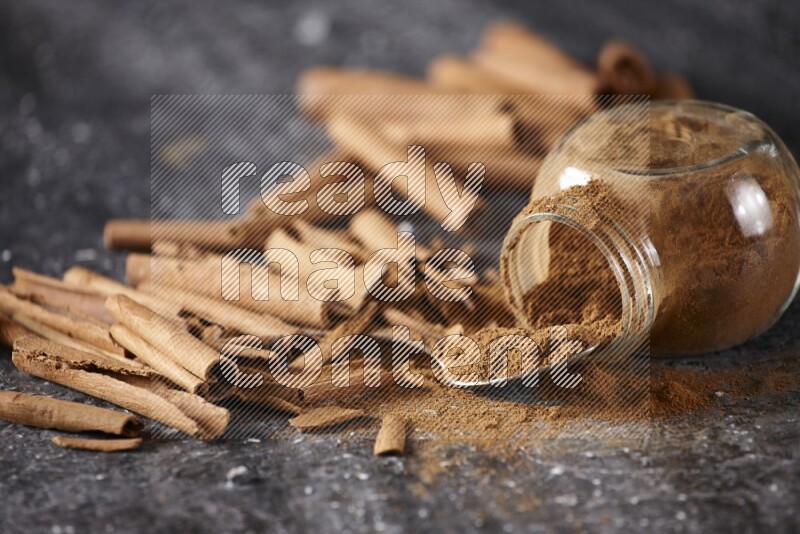 Herbal glass jar full cinnamon powder flipped and a metal spoon full of powder surrounded by cinnamon sticks on textured black background in different angles