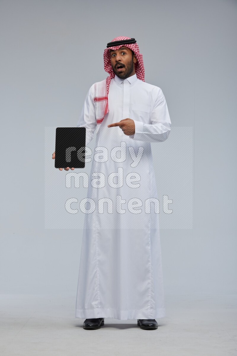 Saudi man Wearing Thob and shomag standing showing tablet to camera on Gray background