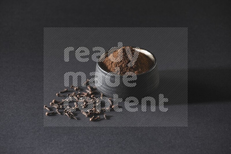 A black pottery bowl full of cloves powder and cloves whole grains on a black flooring
