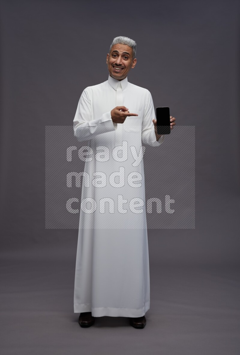 Saudi man wearing thob standing showing phone to camera on gray background