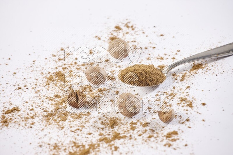 A metal spoon full of nutmeg powder with some of the powder and seeds beside it on a white flooring
