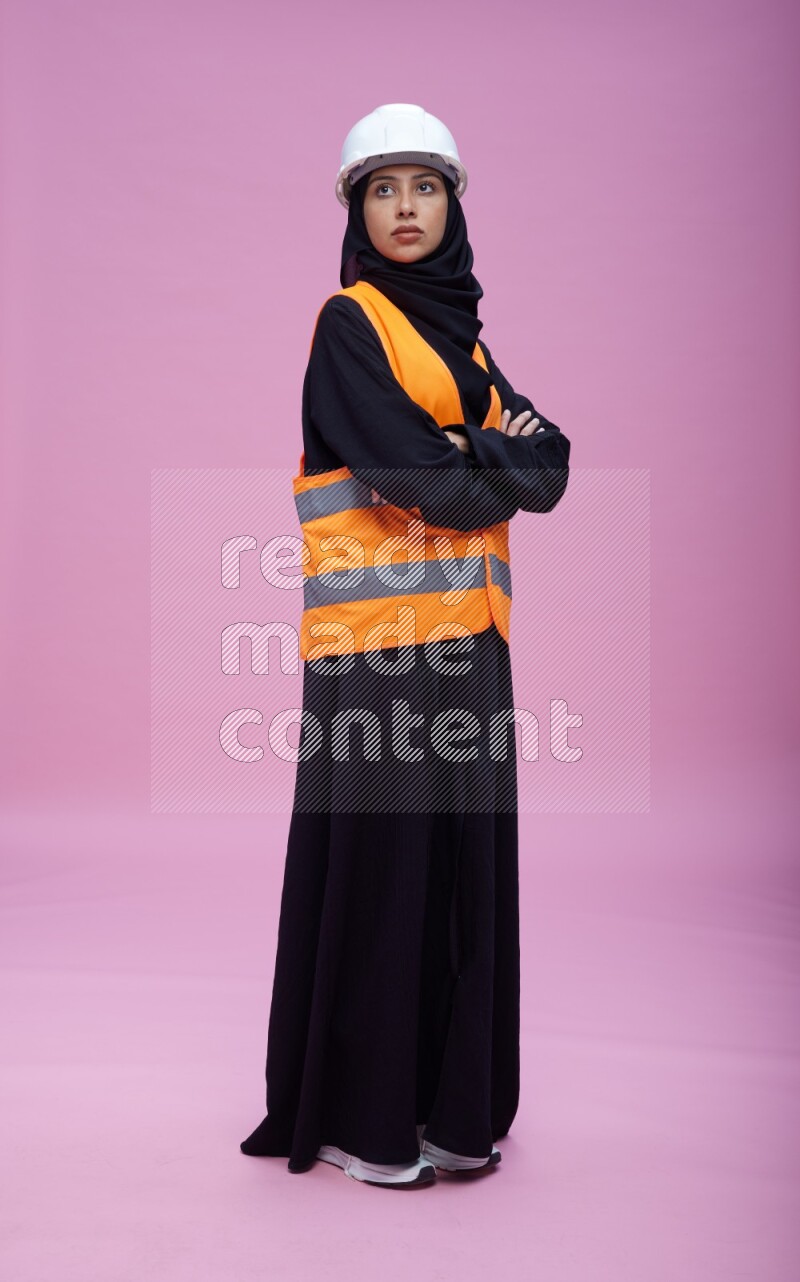 Saudi woman wearing Abaya with engineer vest and helmet standing with crossed arms on pink background