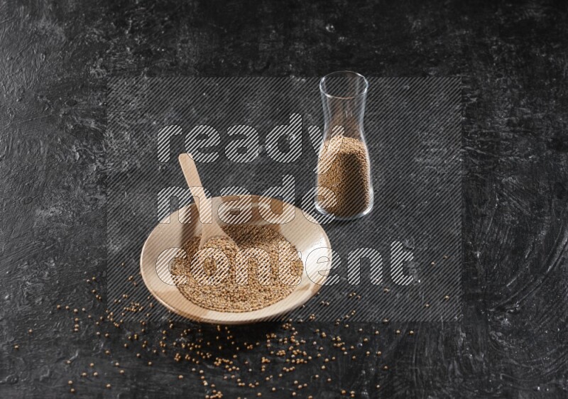 A beige pottery plate full of mustard seeds and a wooden spoon in it with a glass jar filled with the seeds on a textured black flooring