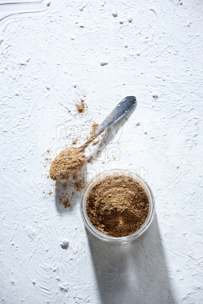 A glass jar and a metal spoon full of allspice powder on a textured white flooring