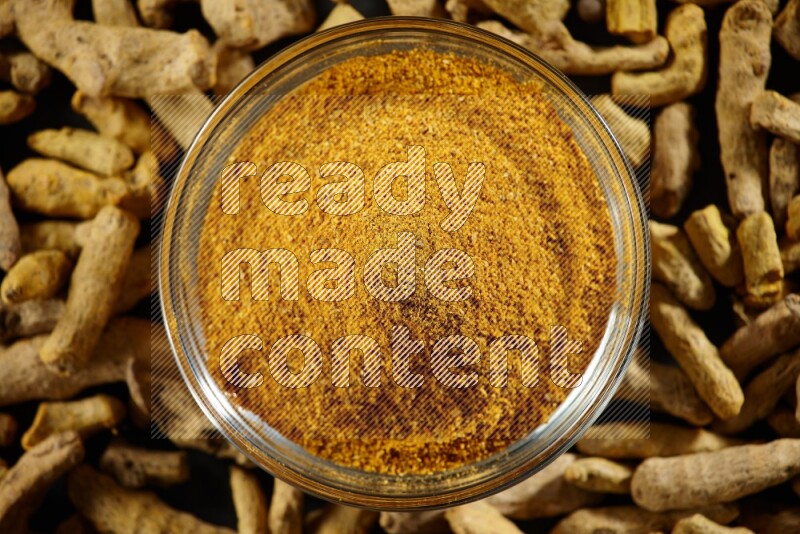 وعاء زجاجي ممتلئ ببودرة الكركم وأصابع الكركم المجففة بجانبه على خلفية سوداء