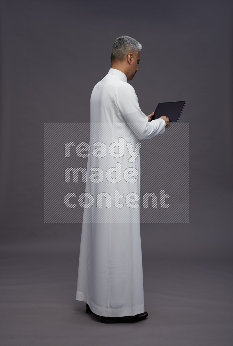 Saudi man wearing thob standing working on tablet on gray background