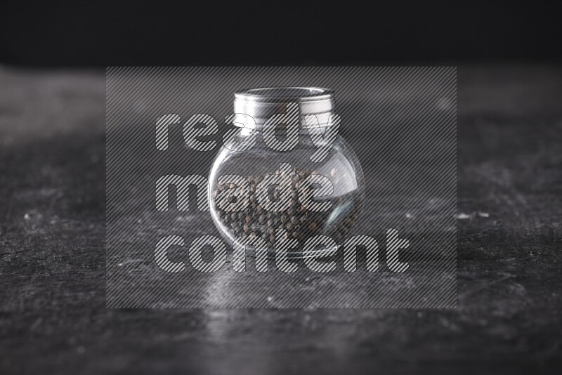A glass spice jar full of black pepper beads on textured black flooring