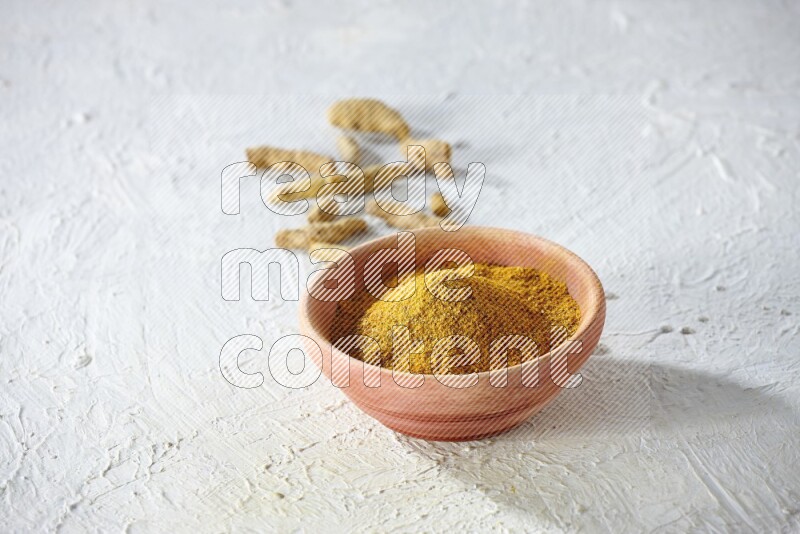 A wooden bowl full of turmeric powder with dried whole fingers on textured white flooring