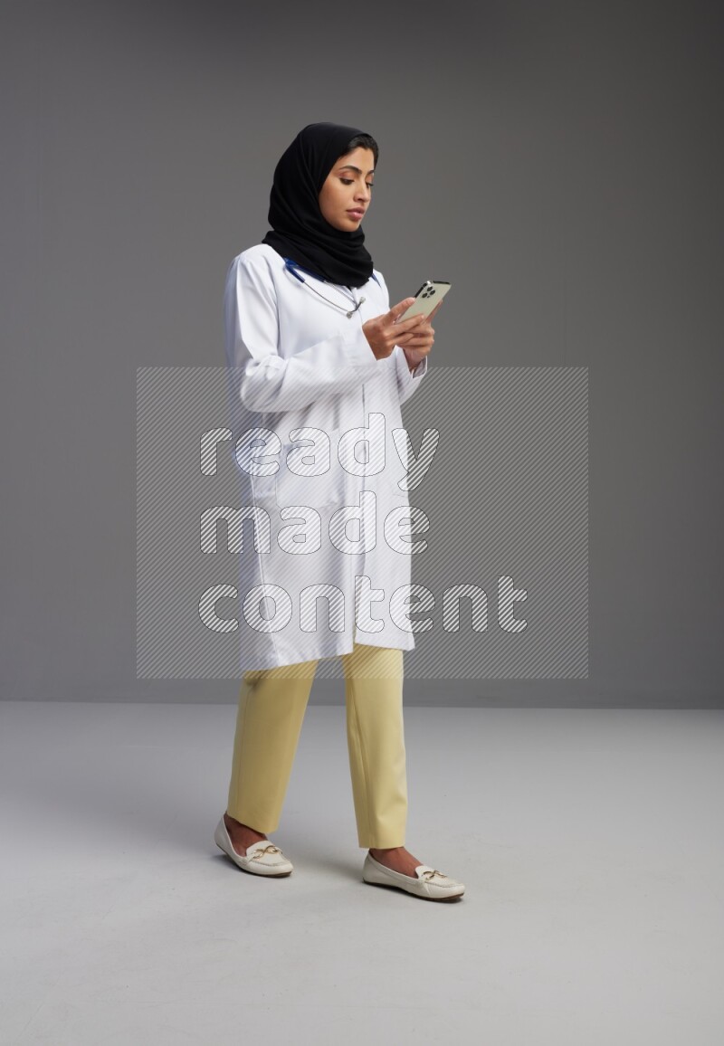 Saudi woman wearing lab coat with stethoscope standing texting on phone on Gray background