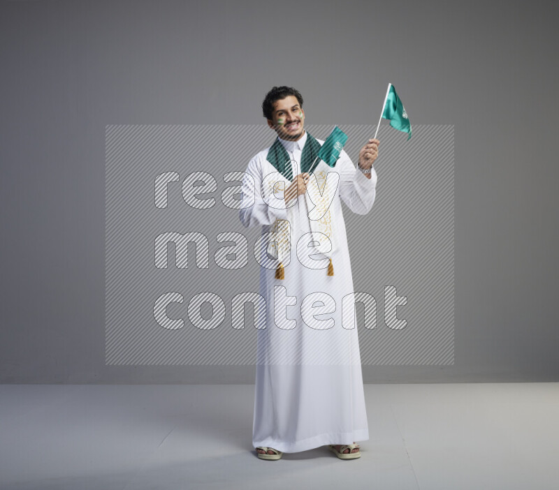 A Saudi man standing wearing thob and saudi flag scarf with face painting holding small Saudi flag on gray background