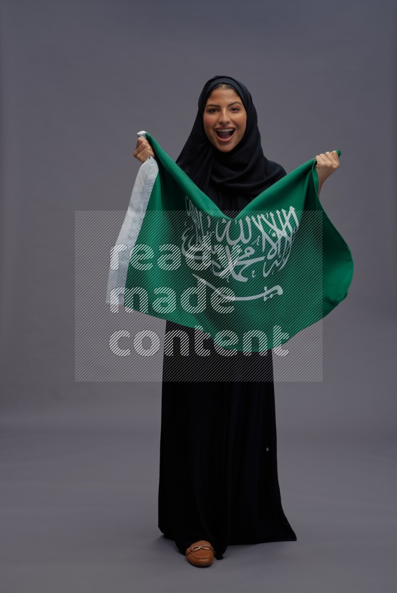 Saudi woman wearing Abaya standing holding Saudi flag on gray background