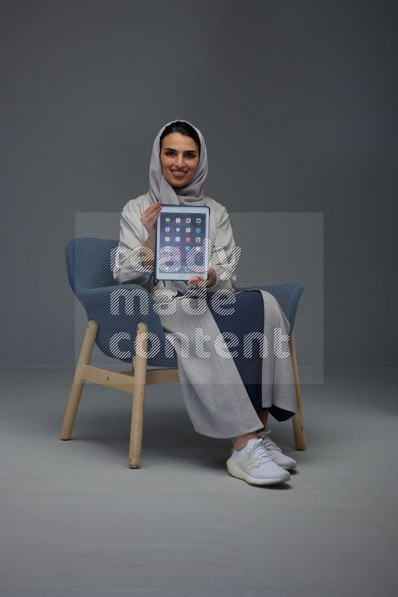A Saudi woman wearing a light gray Abaya and head scarf sitting on a dark grey chair and showing the tablet's screen eye level on a grey background