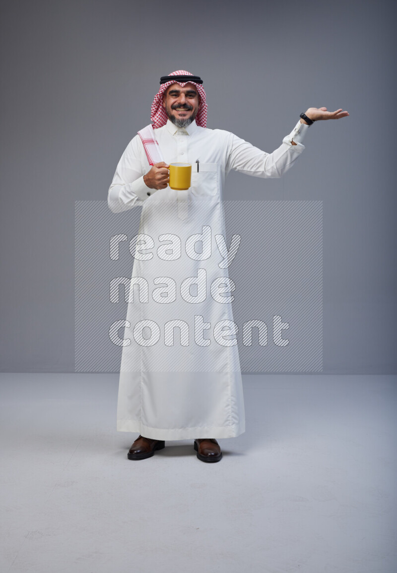 Saudi man Wearing Thob and red Shomag standing holding mug on Gray background